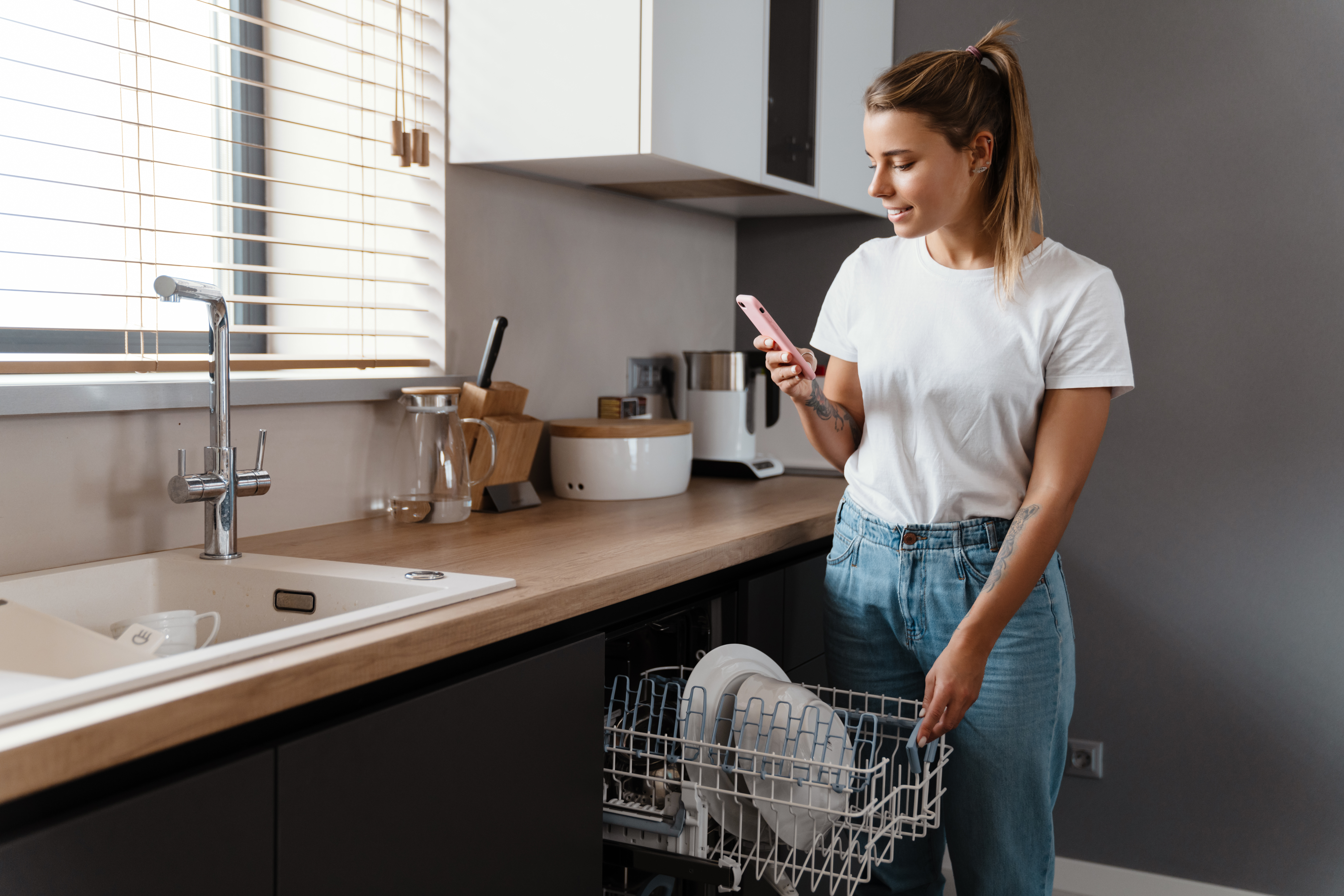 Beautiful happy girl typing on cellphone while using dishwasher